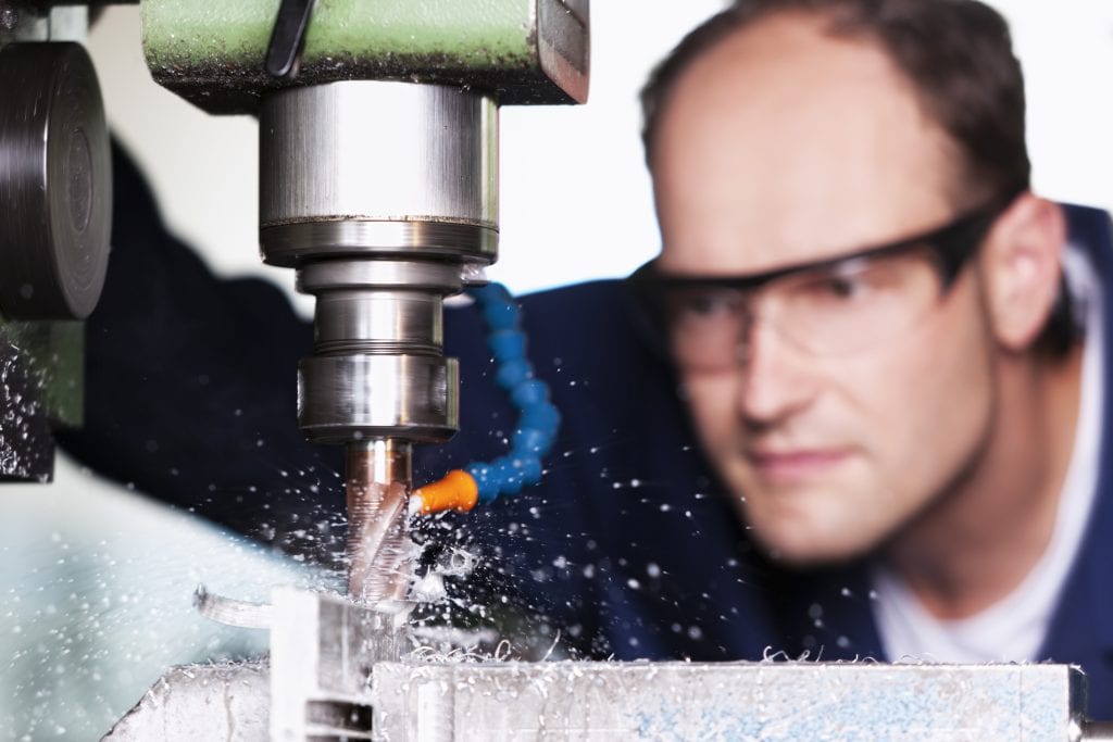 Man wearing safety glasses closely watching milling machine drilling into a block of metal