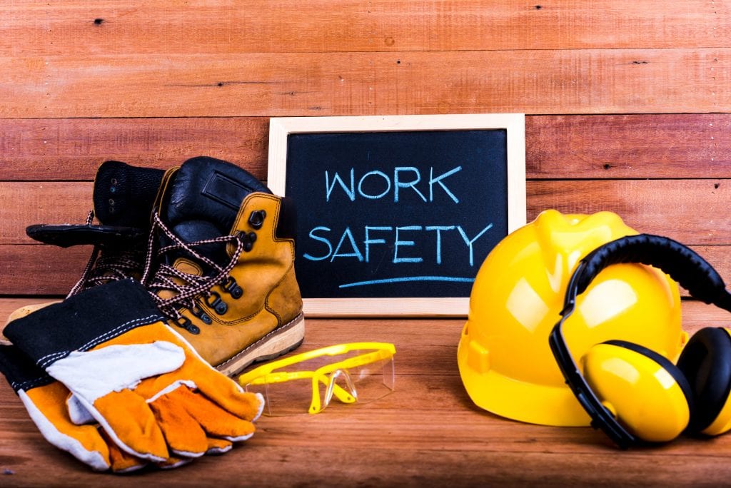 gloves, work boots, safety goggles, hard hat on a wood plank background w/ "work safety" written on a framed chalkboard